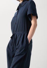 Granatowy kombinezon uszyty z miękkiego materiału, z krótkimi rękawami, zapięciem na guziki z przodu, ściągaczem w pasie oraz bocznymi kieszeniami z haftowanymi detalami.