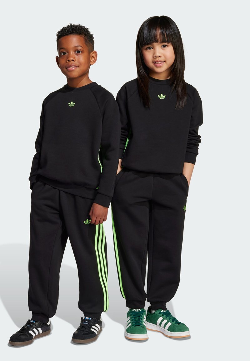 Schwarze sportliche Sweatshirts und Hosen mit neongrünen Seitenstreifen. Logo-Details auf der Brust und dem Oberschenkel. Zwei Kinder posieren nebeneinander.