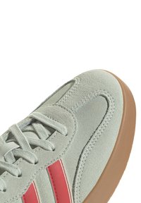 Ljust grå mockasneaker med röda ränder, med gummisula, rund tå och platta snören. Sömmarna framhäver designdetaljerna.