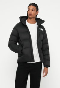 Jeune homme avec des cheveux tressés portant une veste matelassée noire sur une chemise blanche et un pantalon noir, se tenant avec une main dans la poche.
