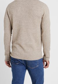 En person i en beige texturerad långärmad tröja och blå jeans, sedd bakifrån mot en enfärgad bakgrund.