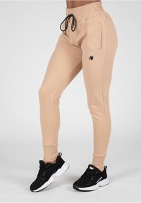 Beige joggers met een elastische tailleband, trekkoord en zijzakken; heeft een zwart logo; gecombineerd met zwarte sportschoenen.