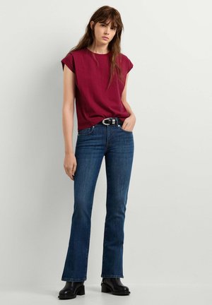 Donna con lunghi capelli castani che indossa una maglia rossa a maniche corte, jeans blu scuro, cintura nera e stivali neri, in piedi contro uno sfondo chiaro e uniforme.