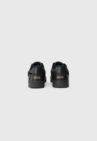 Fekete sportcipők sima bőr és velúr textúrával, párnázott nyakkal, minimalista dizájnnal és arany "VERSACE JEANS COUTURE" logóval a sarkon.