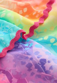 Tissu multicolore à texture lisse, présentant un dégradé de rose, orange, jaune, vert et bleu, bordé d'une bordure festonnée rose.