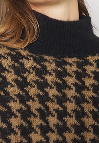 Bruine en zwarte trui met een pied-de-poule patroon en een zwarte geribbelde coltrui, gedragen door een persoon met lichtbruin haar.