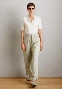 Personne portant un haut blanc à manches courtes à côtes, un pantalon beige ample taille haute, des lunettes de soleil, des boucles d'oreilles créoles et des chaussures beiges marchant sur un sol en bois.