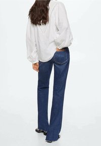 Weiße, strukturierte Bluse mit lockeren Ärmeln und einem V-förmigen Rückendetail, kombiniert mit dunkelblauen, ausgestellten Jeans mit ausgefranstem Saum.