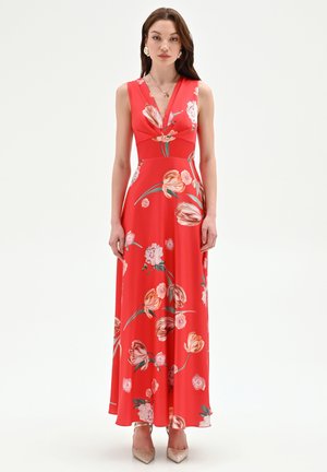 Femme portant une robe longue rouge sans manches avec imprimé floral rose et décolleté en V, debout devant un fond clair uni.