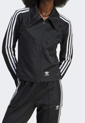 Person trägt einen schwarzen Adidas-Trainingsanzug mit weißen Streifen an Ärmeln und Hosen, Reißverschlussjacke und Adidas-Logo auf Brust und Oberschenkel.