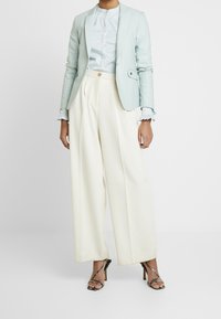 Ljust blå skräddarsydd kavaj över en mjuk, vit blus och vida, krämfärgade byxor. Svarta remsandaler kompletterar outfiten.