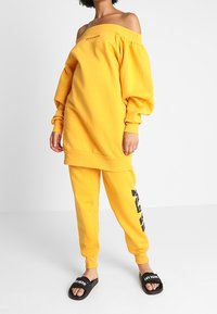 Sudadera amarilla de gran tamaño, con los hombros al aire y mangas abullonadas, pantalones jogger amarillos a juego con acentos gráficos negros y sandalias negras tipo slide.
