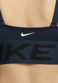 Zadnji pogled na osebo, ki nosi temno modre Nike športne nedrčke z nastavljivimi naramnicami in črnimi črkami "NIKE" čez pas.