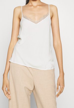 Kvinde iført hvid camisole med blondestrimle og beige vidde benbukser, vist fra nakke til midt på låret mod en ensfarvet baggrund.