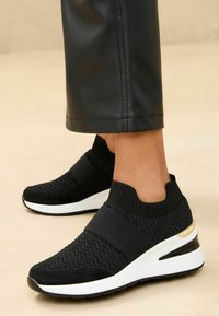 Baskets en tricot noires avec une tige texturée, une sangle élastique et une semelle intermédiaire blanche élégante avec un accent doré au talon.