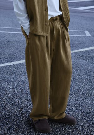 Osoba ubrana w luźne oliwkowo-brązowe spodnie ze ściągaczem w talii i dopasowaną kamizelkę, białą koszulę z długim rękawem oraz ciemnobrązowe buty, stojąca na chodniku.