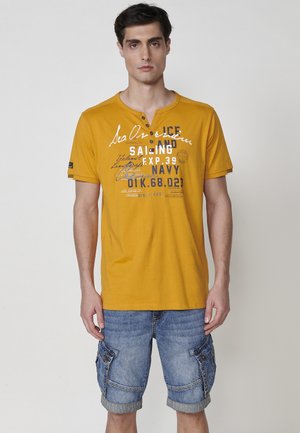 Camiseta amarilla de manga corta con escote en V, que presenta un texto impreso en blanco y azul. Combinada con pantalones cortos de mezclilla tipo cargo, con bolsillos.