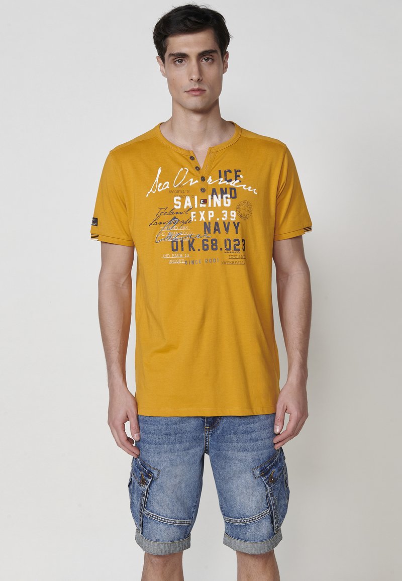 Trumpa rankovėmis geltona marškinėlė su v-kaklu, papuošta balta ir mėlyna spausdinta tekstu. Derinama su džinsiniais šortais su kišenėmis.