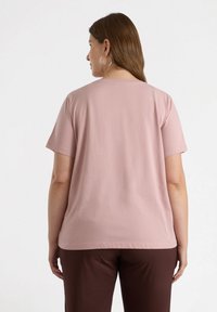 Camiseta de manga corta de color rosa claro con cuello redondo, textura suave y ajuste relajado, combinada con pantalones marrones. Sin patrones ni acentos visibles.
