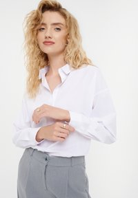 White button-up shirt with a collar, made from smooth fabric, paired with gray trousers featuring a single button closure and tailored fit.
