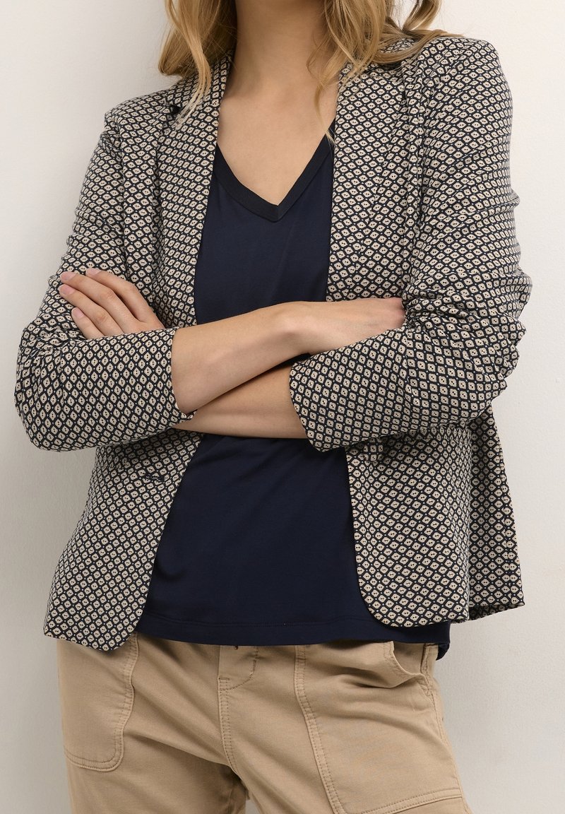 Personne les bras croisés portant un blazer à motifs, une chemise bleu marine à col en V et un pantalon beige sur un fond clair uni.