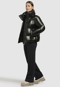 Doudoune noire avec une texture brillante et matelassée et un col haut. Portée par-dessus un haut noir, associée à un pantalon noir et des baskets compensées beiges.
