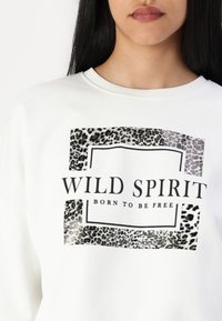 Witte sweatshirt met een luipaardprint afbeelding en de tekst "WILD SPIRIT BORN TO BE FREE" in bold zwarte letters, ronde halslijn.