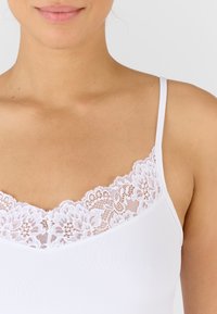 Witte camsol met een geschulpte kantafwerking rond de halslijn, een gladde textuur en verstelbare dunne bandjes.