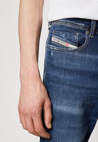 Vaqueros de mezclilla azul oscuro con parches desgastados. Cuenta con un diseño de cinco bolsillos y una etiqueta de marca visible en el bolsillo trasero.