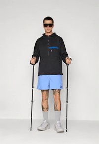 Μαύρος φλις πόλο με μπλε θηκάκι με φερμουάρ, συνδυασμένο με ανοιχτό μπλε σορτσάκι. Το άτομο κρατάει μιζέτες και φοράει γκρι κάλτσες και αθλητικά παπούτσια.