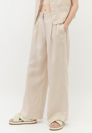 Osoba nosząca luźne, beżowe spodnie z wysokim stanem i szerokimi nogawkami z bocznymi kieszeniami oraz jasnzielone sandały na krzyż, stojąca na jednolitym tle.
