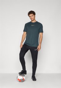 Nike sporta apģērbs, kas sastāv no zilo īso piedurknju krekla un melnām biksēm, ar izteiktu logotipu. Novietots blakus futbola bumbai.
