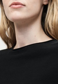 Gros plan sur le cou et le bas du visage d'une personne à la peau claire, aux cheveux châtain clair, portant un haut noir texturé.