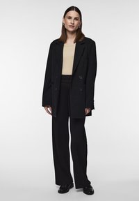 Blazer noir à double boutonnage sur un top beige, associé à un pantalon plissé noir et des chaussures noires brillantes, avec un design simple et élégant.