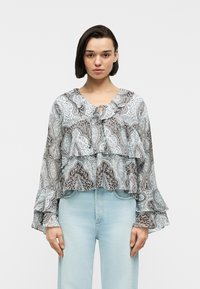 Femme portant un chemisier transparent à motifs avec des manches et un col à volants, associé à un jean taille haute bleu clair, debout devant un fond blanc.