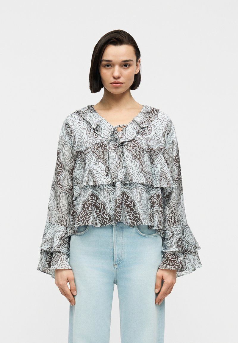 Femme portant un chemisier transparent à motifs avec des manches et un col  à volants, associé à un jean taille haute bleu clair, debout devant un fond blanc.