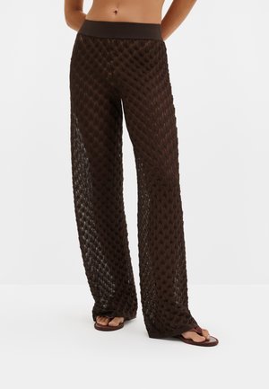 Pantalon large marron en maille texturée porté avec des sandales marron à bout ouvert, taille du modèle visible, sur un fond clair uni.