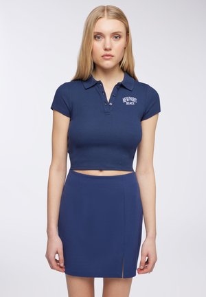 Polo cropped blu navy con maniche corte e colletto, con logo di Newport Beach. Abbinato a una gonna alta coordinata, con un leggero dettaglio di spacco.