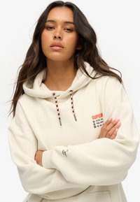 Haut à capuche couleur crème avec une poche avant, cordons de serrage contrastants noirs et orange, et logo imprimé sur la poitrine et la manche.