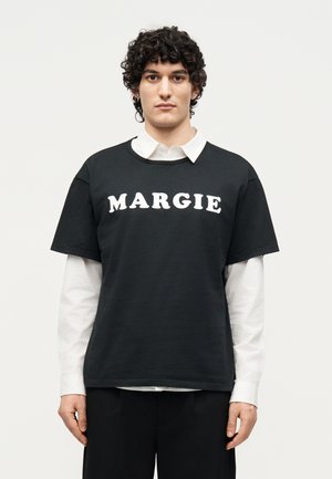 Jeune homme aux cheveux bouclés portant une chemise blanche à col superposée et un t-shirt noir avec le texte « MARGIE » à l'avant, debout devant un fond uni.