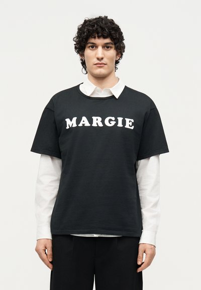 Joven con cabello rizado vistiendo una camisa blanca con cuello en capas y una camiseta negra con el texto "MARGIE" en el frente, de pie sobre un fondo liso.