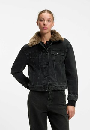 Frau trägt eine schwarze Jeansjacke mit braunem Fellkragen und passenden schwarzen Jeans, steht mit einer Hand in der Hüfte vor einem weißen Hintergrund.
