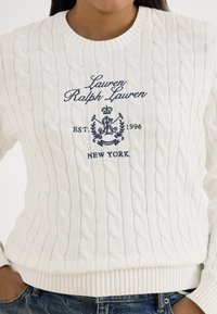 Biały sweter z dzianiny warkoczowej z granatowym napisem "Lauren Ralph Lauren EST. 1996 NEW YORK" i herbem wyszywanymi na piersi, zestawiony z niebieskimi dżinsami.