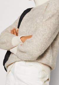 Beiger Strickpullover mit strukturiertem Finish, ergänzt durch gerippte Bündchen und einen hohen Kragen, kombiniert mit weißen Hosen und einem schwarzen Gurt.