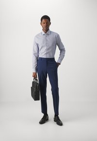 Camicia a righe azzurro chiaro, pantaloni navy su misura, scarpe nere, che tiene una borsa in pelle nera, in piedi su uno sfondo grigio minimalista.