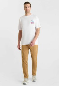 Man in een wit graphic t-shirt met de tekst "Let op, Boze Baas," beige broek met grote voorzakken en grijze sneakers, staand op een witte achtergrond.