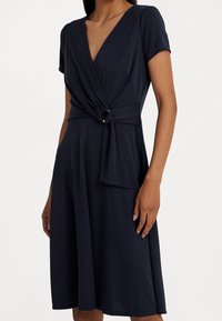 Dress navy a maniche corte con scollatura a V, dettaglio a torsione frontale e accentuazione circolare in vita, realizzato in tessuto liscio con una silhouette fluida.
