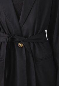 Schwarzer Wickelblazer aus strukturiertem Stoff mit einem um die Taille gebundenen Gürtel und einem runden goldenen Knopf. Verfügt über zwei Seitentaschen.