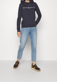 Granatowy hoodie z białym logo "GANT 1949", połączony z jasnoniebieskimi dżinsami i ciemnymi trampkami z żółtymi akcentami oraz teksturowanymi podeszwami.