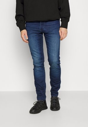 Osoba ubrana w ciemnoniebieskie dopasowane jeansy, czarne trampki z ozdobnymi sznurówkami oraz czarną bluzkę z długim rękawem, stojąca na jednolitym tle.